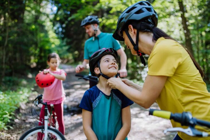 Jovem família, com quatro elementos (duas crianças e dois adultos) param de andar de bicicleta, enquanto a mãe ajeita o capacete ao filho mais velho.