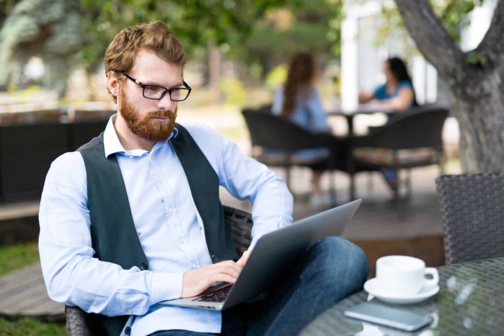 Homem ruivo de barba e óculos trabalha numa esplanada, com o computador portátil e um café na mesa à sua frente.