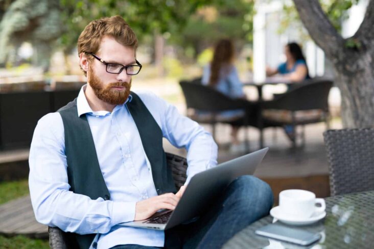 Homem ruivo de barba e óculos trabalha numa esplanada, com o computador portátil e um café na mesa à sua frente.