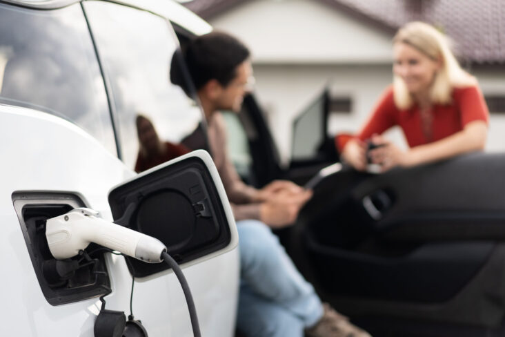 duas pessoas a conversar uma fora do carro e outra sentada no carro com a porta aberta a ilustrar os apoios à compra de um carro elétrico.