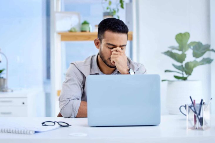 Jovem preocupado diante do computador portátil, com óculos em cima da secretária.