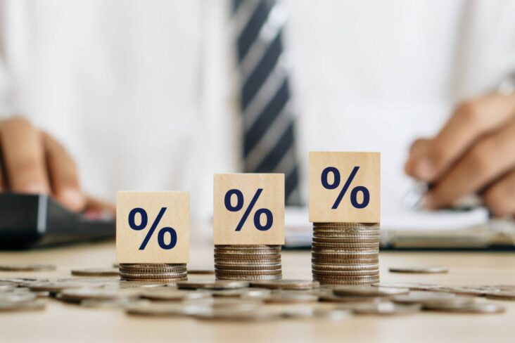 três montes de moedas de euro ao lado uns dos outros. Ao cimo de cada um estão três cubos de madeira com o símbolo de percentagem, numa alusão aos juros, taxas e tarifas