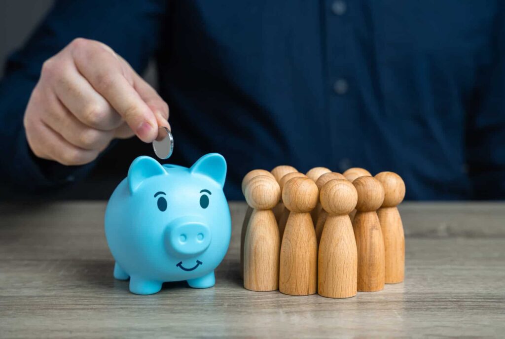 O que é um fundo PPR e quais as vantagens? Um homem coloca uma moeda num porco-mealheiro azul (com cara virada para a câmara) com peões de madeira ao lado, tudo em cima de uma mesa.