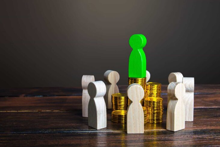 Peças em madeira, a representar pessoas, formam uma roda em torno de alguns montes de moedas, numa alusão à poupança e ao investimento