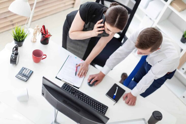 Dois funcionários de uma empresa trabalham lado a lado, confirmando informações num computador, enquanto um deles está ao telefone.