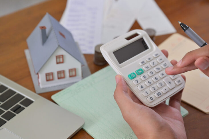 mãos a usar uma calculadora, sobre a mesa há um computador, papéis e uma casa em miniatura - para ilustrar artigo sobre "Venda da casa: Como sei quanto vale a minha casa"