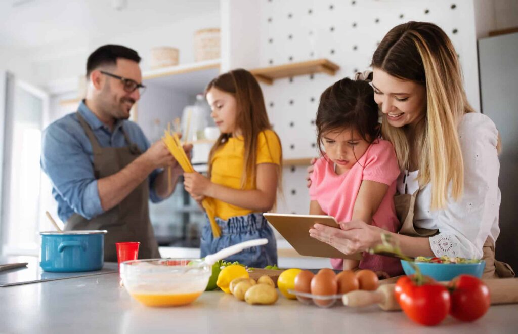 Família de quatro elementos (dois adultos e duas crianças) estão na cozinha a preparar uma refeição em conjunto