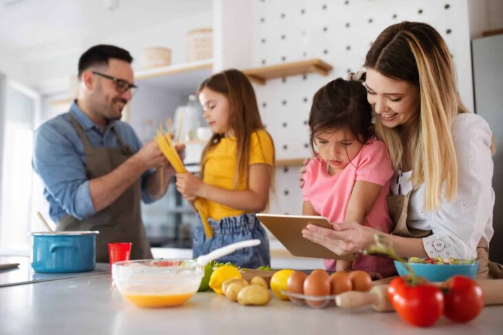 Família de quatro elementos (dois adultos e duas crianças) estão na cozinha a preparar uma refeição em conjunto