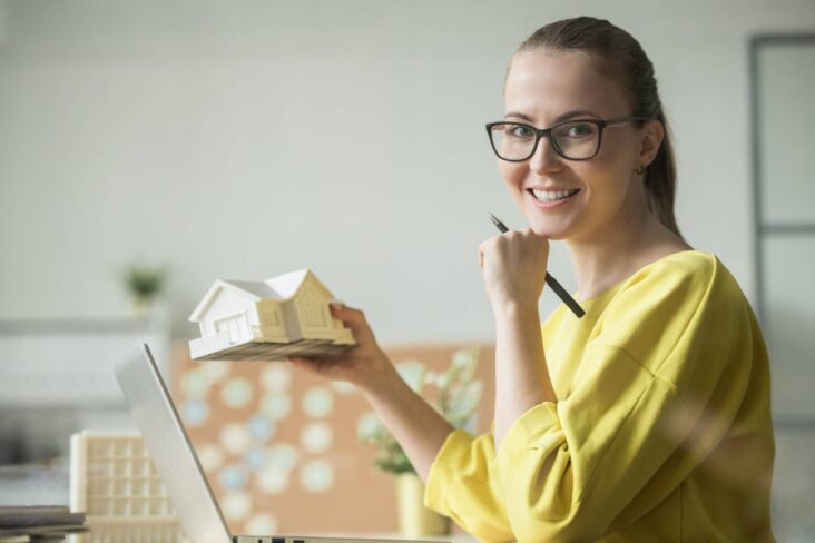 jovem mulher freelancer frente ao computador a segurar uma casa na mão a ilustrar o tema de como comprar casa como freelancer