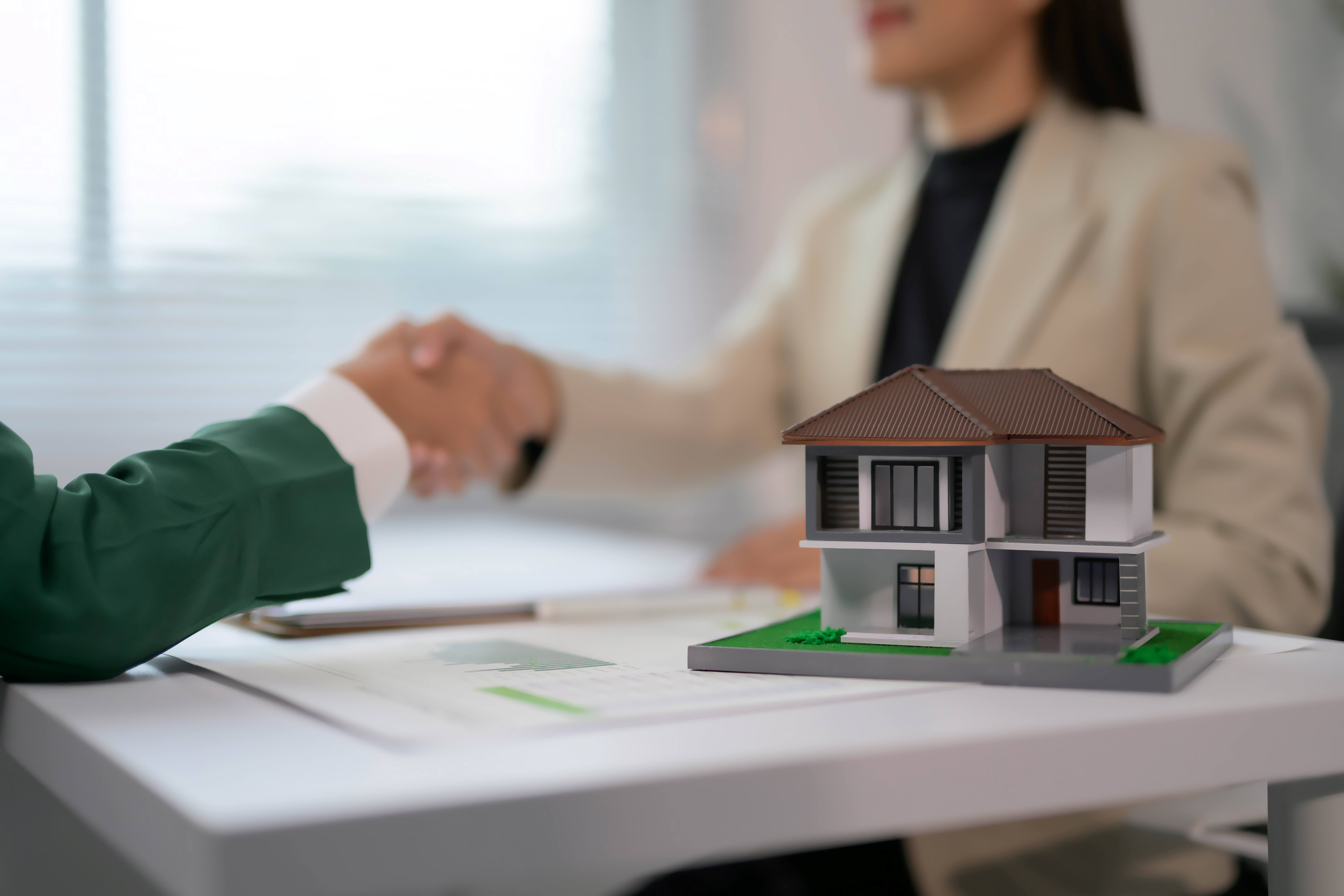 Agente imobiliário apertando a mão do cliente sobre modelo de casa na mesa em um ambiente de escritório moderno