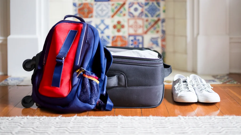 Entrou numa universidade longe de casa? Este é o seu guia de sobrevivência Mochila encostada a mala de viagem aberta, ao lado de umas sapatilhas por calçar