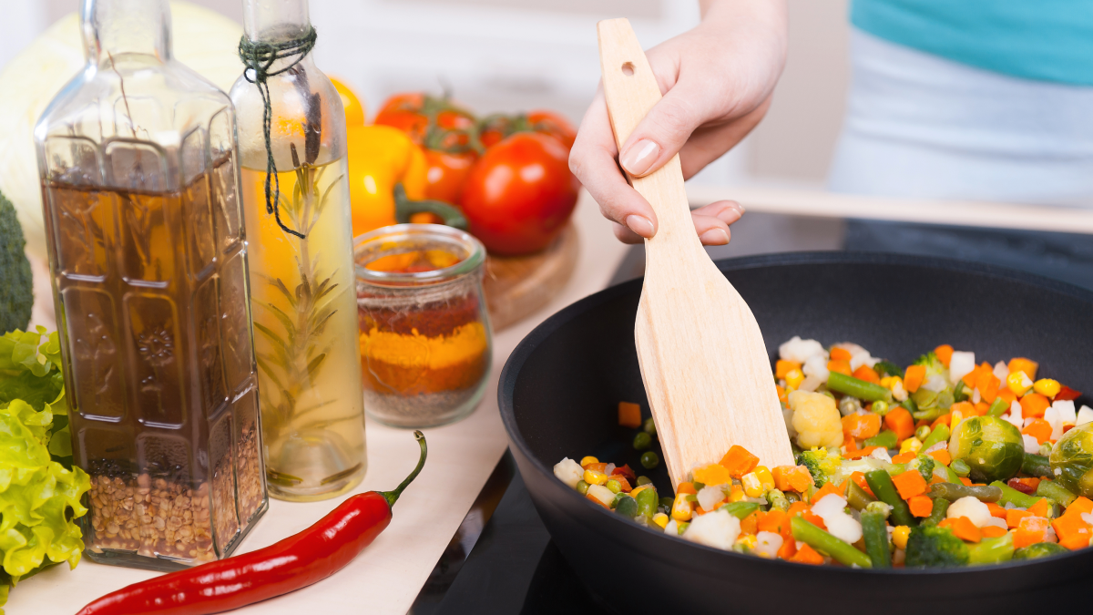Pessoa cozinha legumes numa frigideira ao lado de vários condimentos e vegetais