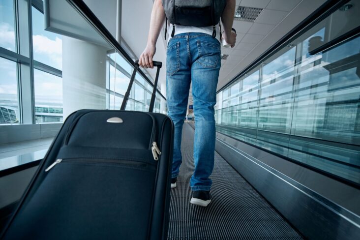imagem de um homem de costas num tapete rolante no areoporto com uma mala de viagem - para ilustar artigo sobre seguros de viagem para quem vai de férias