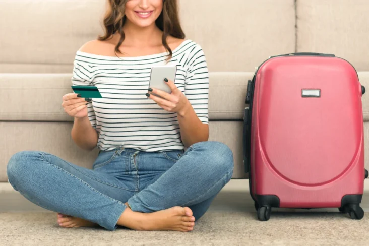 mulher sentada no chão com uma mala de viagem ao lado e com um cartão de crédito e o telemóvel na mão a ilustrar as vantagens dos cartões de crédito com milhas