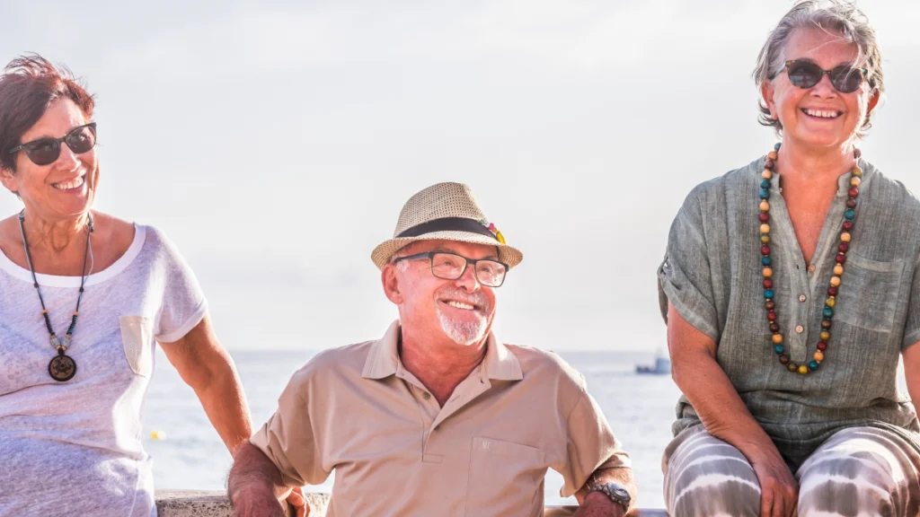 Três pessoas, aparentemente reformadas, sentadas à beira mar, a aproveitar a vida