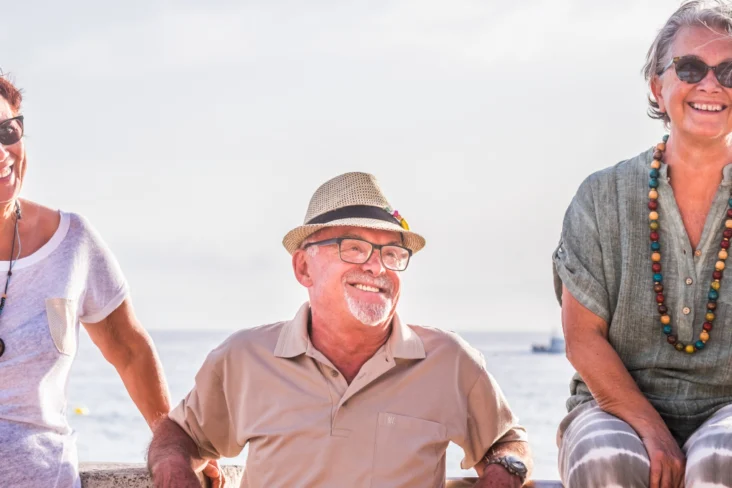 Três pessoas, aparentemente reformadas, sentadas à beira mar, a aproveitar a vida