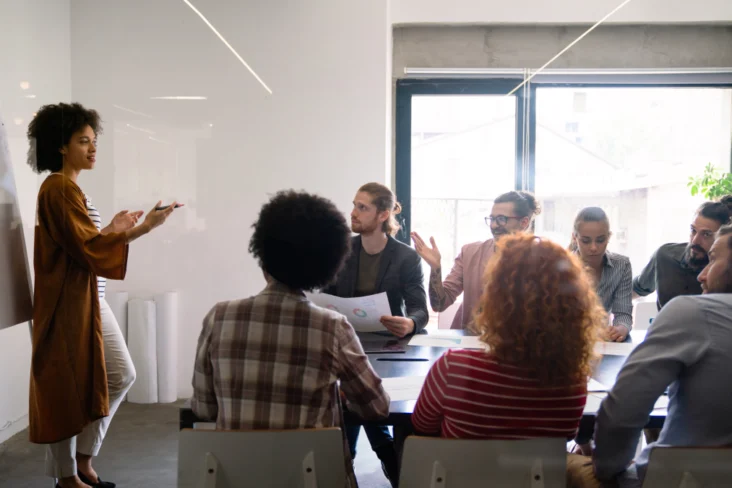 Mulher dá uma formação a um grupo de sete pessoas sentadas à volta de uma mesa de escritório