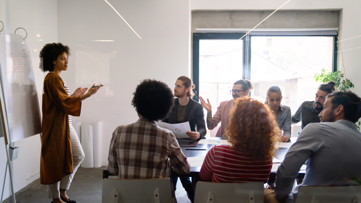 Mulher dá uma formação a um grupo de sete pessoas sentadas à volta de uma mesa de escritório