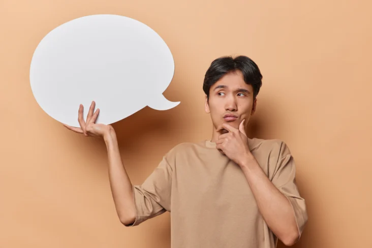 Homem segura num balão de fala feito de papel, enquanto posa com uma careta pensativa